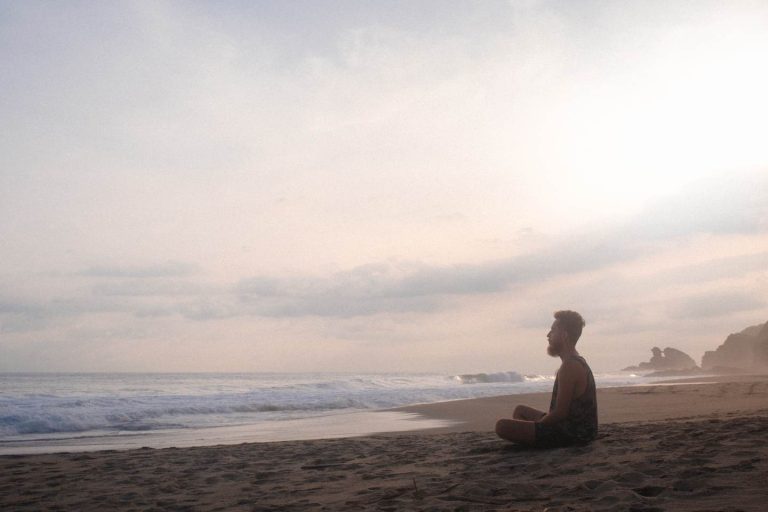 Ein Mann sitzt mit geschlossenen Augen am Strand im Schneidersitz und meditiert - Meditieren zu lernen zahlt sich aus.